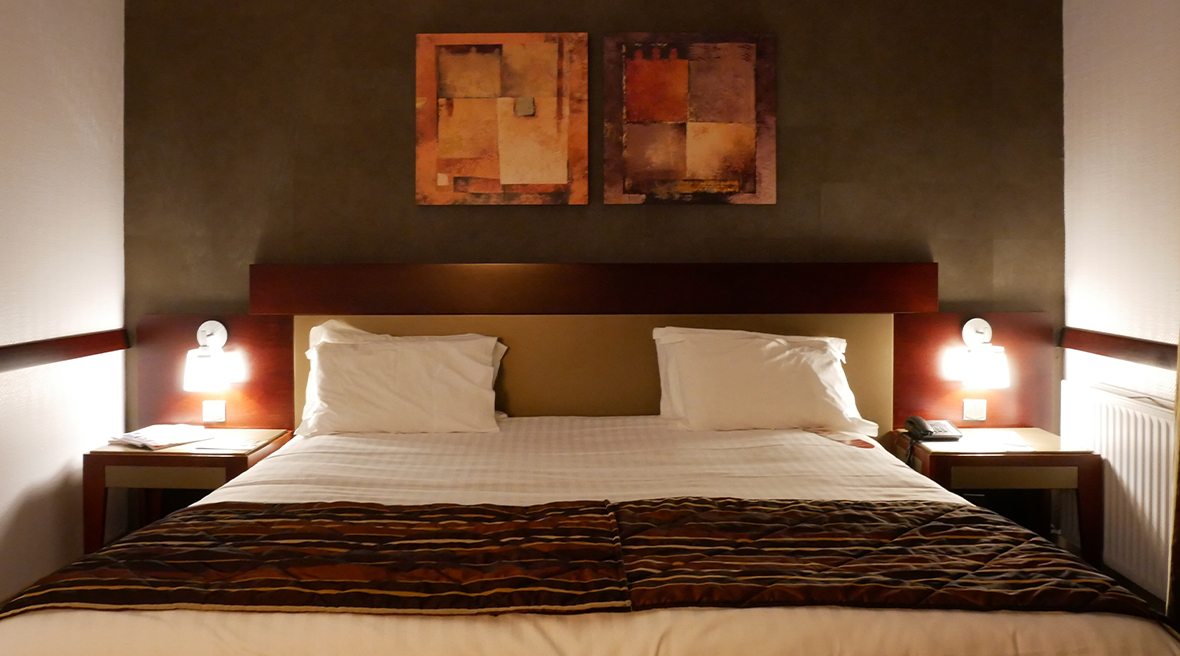 An inviting double bed against a dark brown wall with bedside lamps either side and two pieces of art hanging above the bed frame